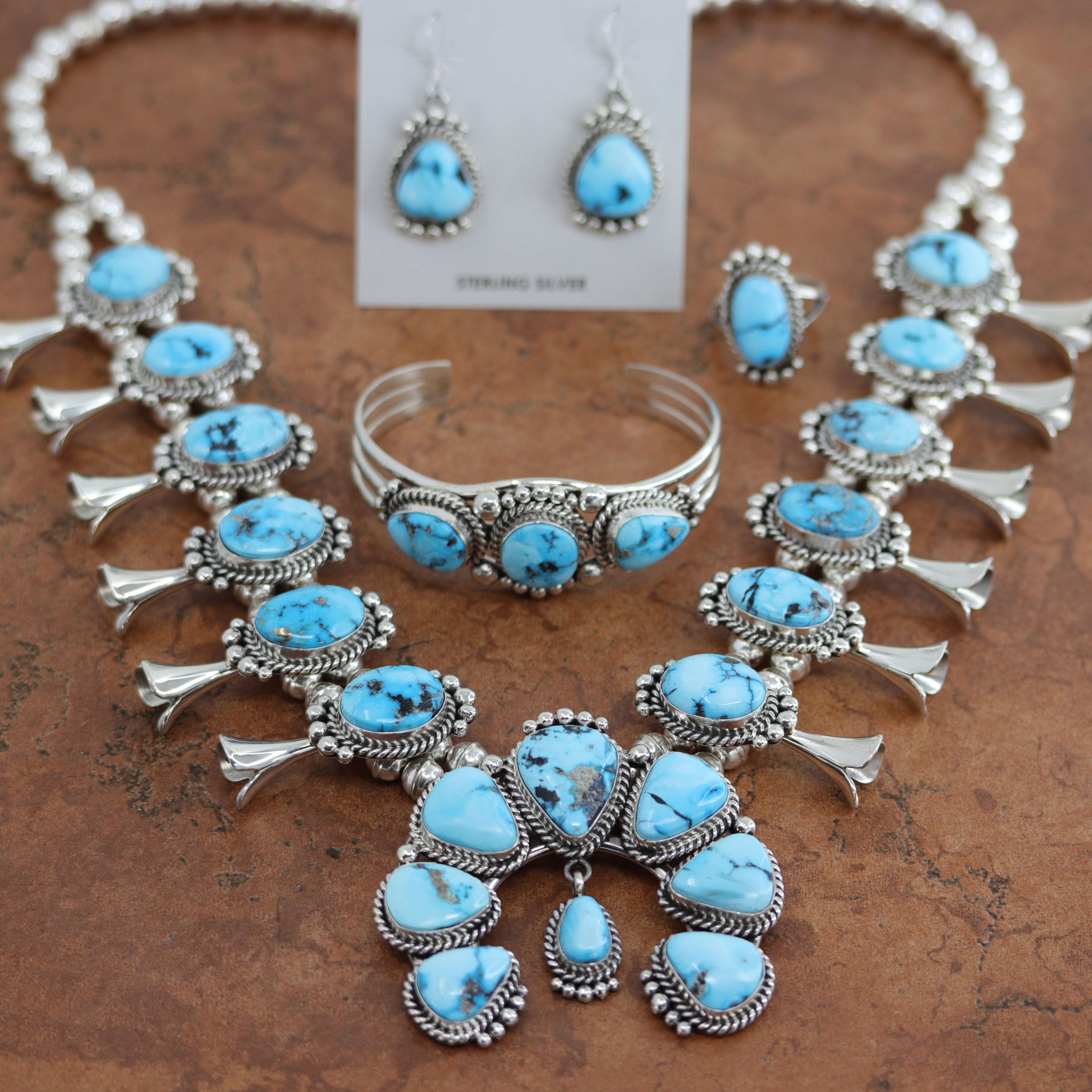 An authentic Navajo Squash Blossom set including necklace, earrings, bracelet, and ring. It is made from sterling silver and turquoise. The necklace is about 26". The Naja is 2 7/8" by 2 3/4". Each blossom is 1 7/8" in length. All the pieces are stamped by the artist and Sterling. The bracelet is 5 3/4" around and 1" in width. The opening on it is 1 1/8" and it is partly flexible. The earrings are 1 5/8" in length including the hook and 5/8" in width. The ring is a size 8 and it is 1" in length.