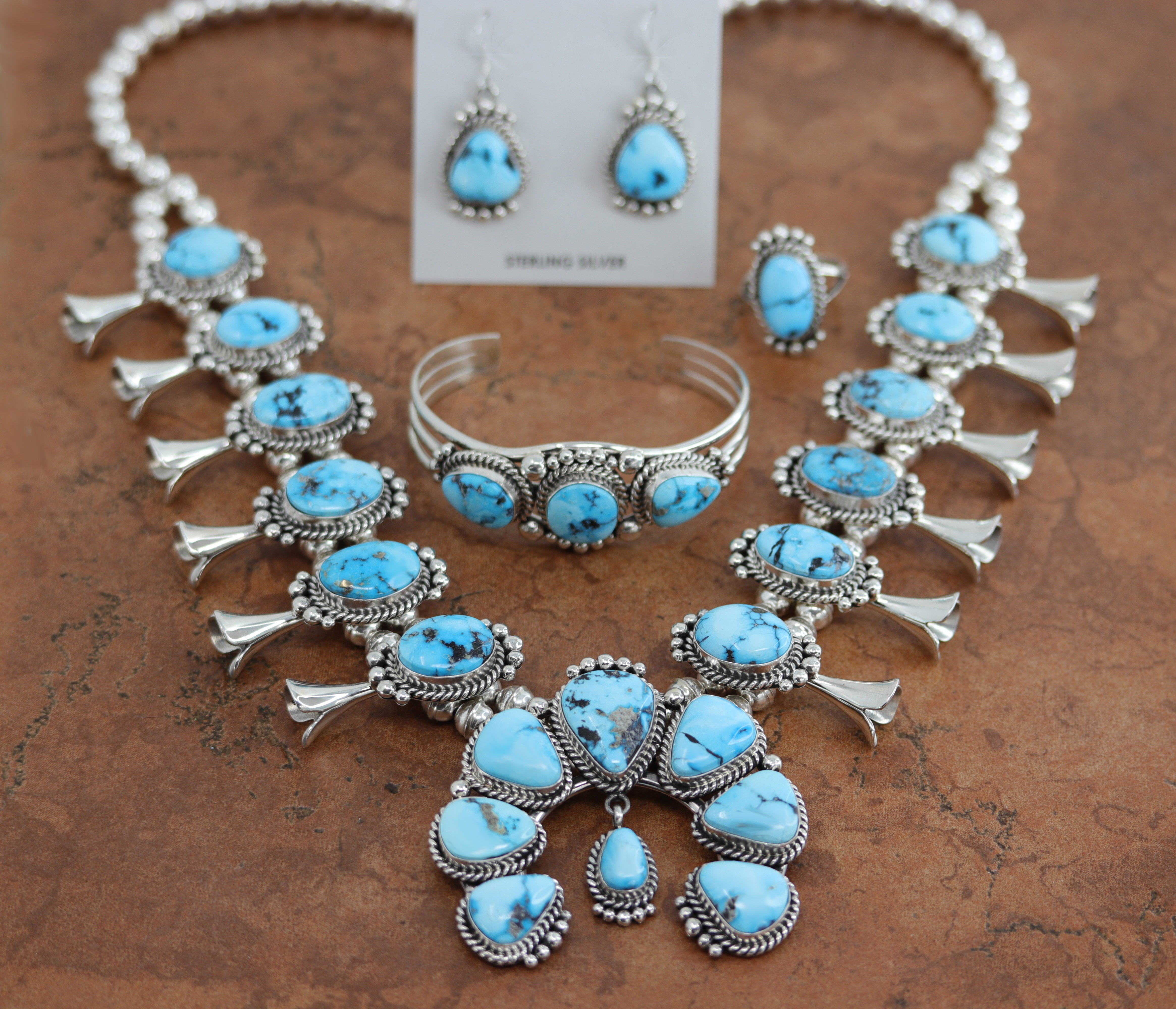 An authentic Navajo Squash Blossom set including necklace, earrings, bracelet, and ring. It is made from sterling silver and turquoise. The necklace is about 26". The Naja is 2 7/8" by 2 3/4". Each blossom is 1 7/8" in length. All the pieces are stamped by the artist and Sterling. The bracelet is 5 3/4" around and 1" in width. The opening on it is 1 1/8" and it is partly flexible. The earrings are 1 5/8" in length including the hook and 5/8" in width. The ring is a size 8 and it is 1" in length.